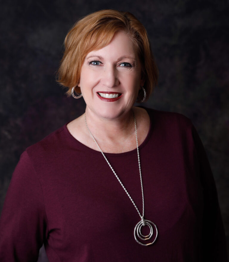 Studio portrait of Elizabeth Toller smiling against a dark background, wearing a burgundy top and silver necklace in soft, controlled light.
